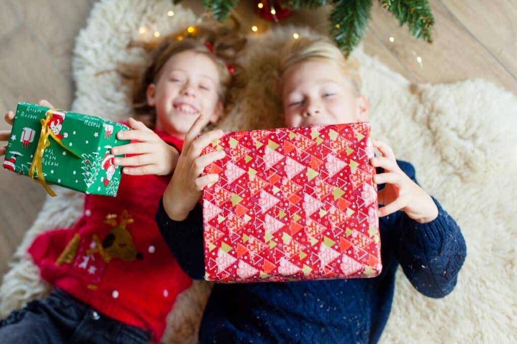 How to Christmas Shop Under $50 Per Kid (in Total) featured image - shows two children laying on the ground holding Christmas presents
