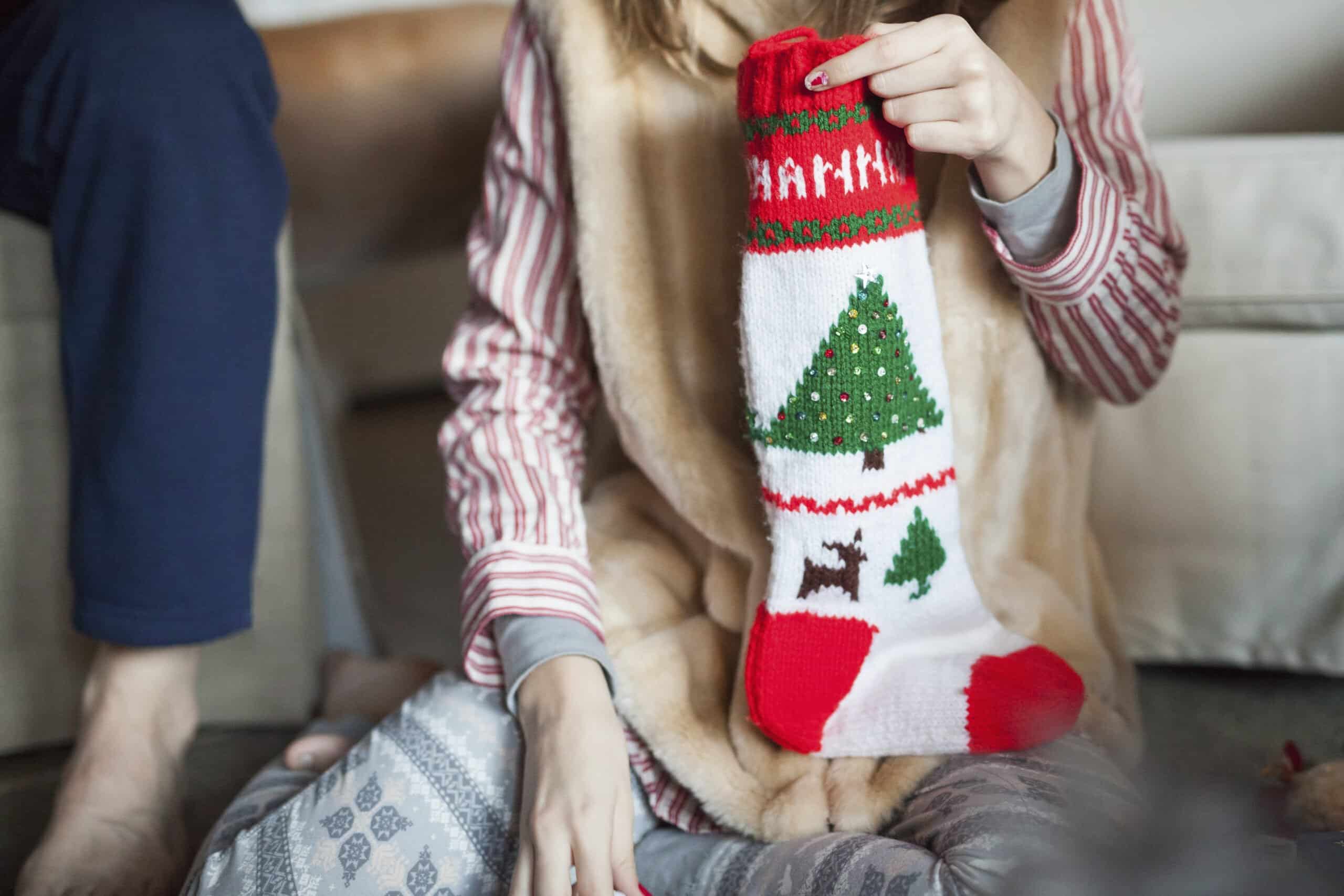 Featured image for Stocking Stuffer Ideas for Mom - shows a woman in pyjamas holding a Christmas stocking